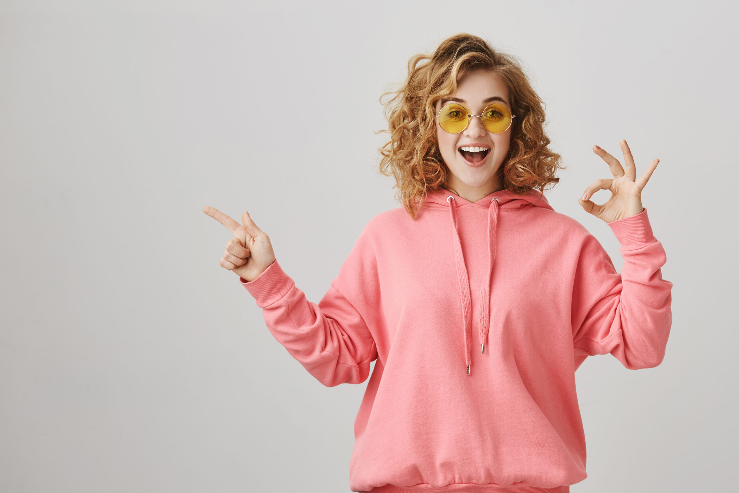 Studio portrait of emotive happy feminine woman with curly hair, wearing casual hoodie and sunglasses showing ok sign and pointing left with index finger, smiling with excitement over gray background.