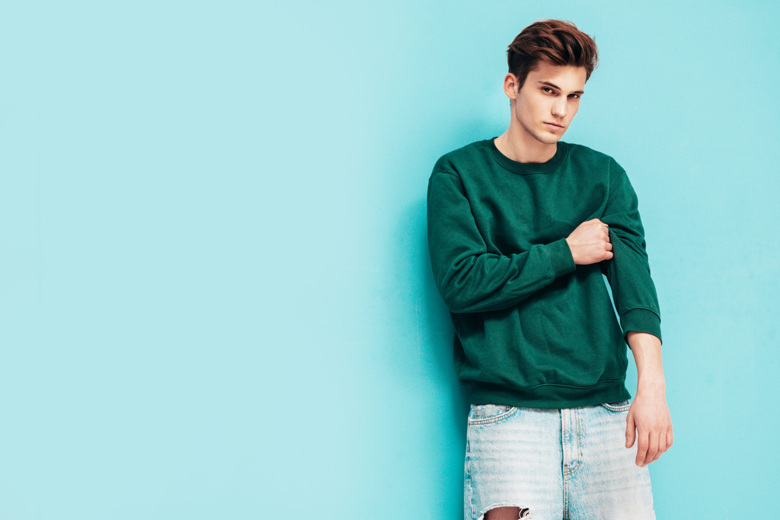 Portrait of handsome confident stylish hipster lambersexual model.Man dressed in green sweater and jeans. Fashion male posing in studio near blue wall